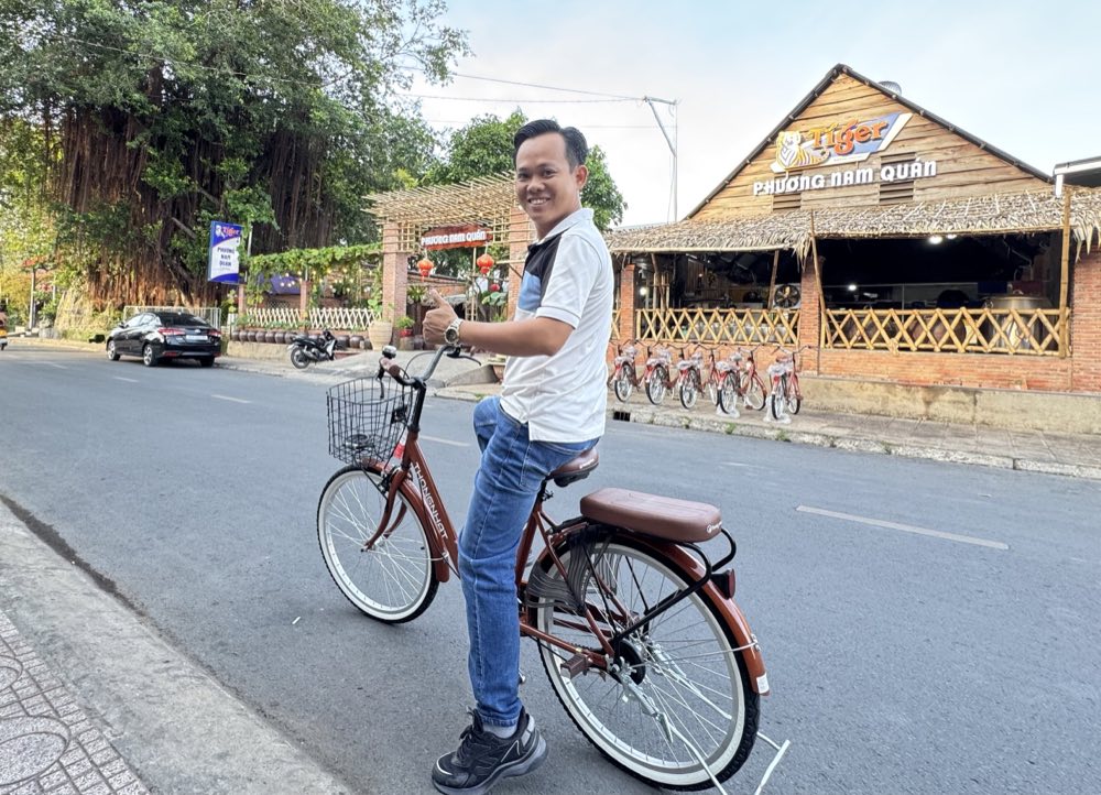 Nguyễn Tấn Lực trên xe đạp trước Phương Nam Quán, nhà hàng mái lá bên đường